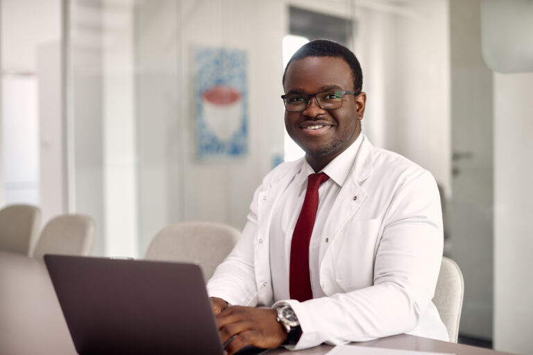 Happy African American Doctor Working On Laptop And The Clinic And Looking At Camera. - ADF PRO