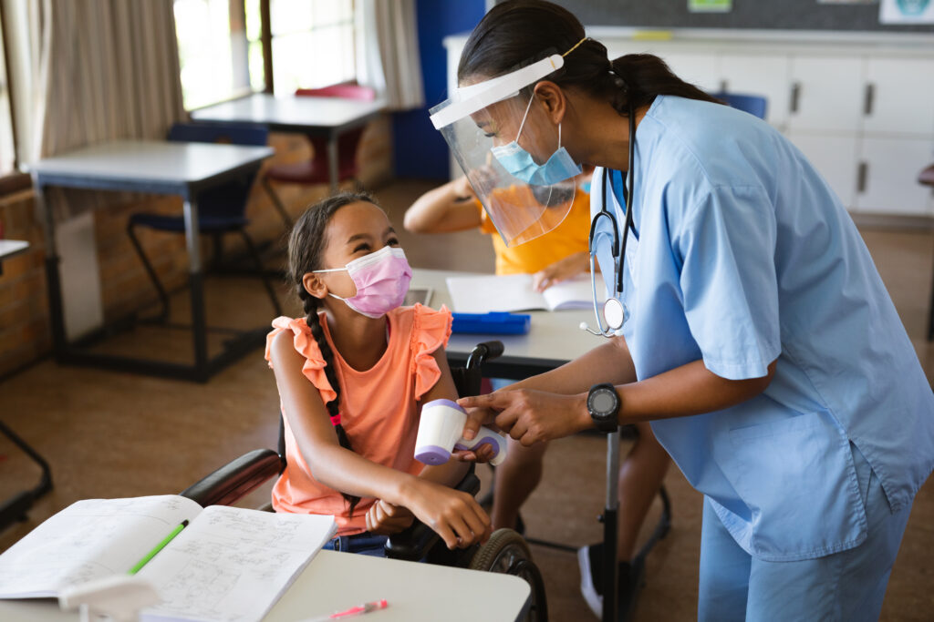 Female Health Worker Wearing Face Shield Measuring Temperature Of Disabled Girl At Elementary School. Education Back To School Health Safety During Covid19 Coronavirus Pandemic. - ADF PRO